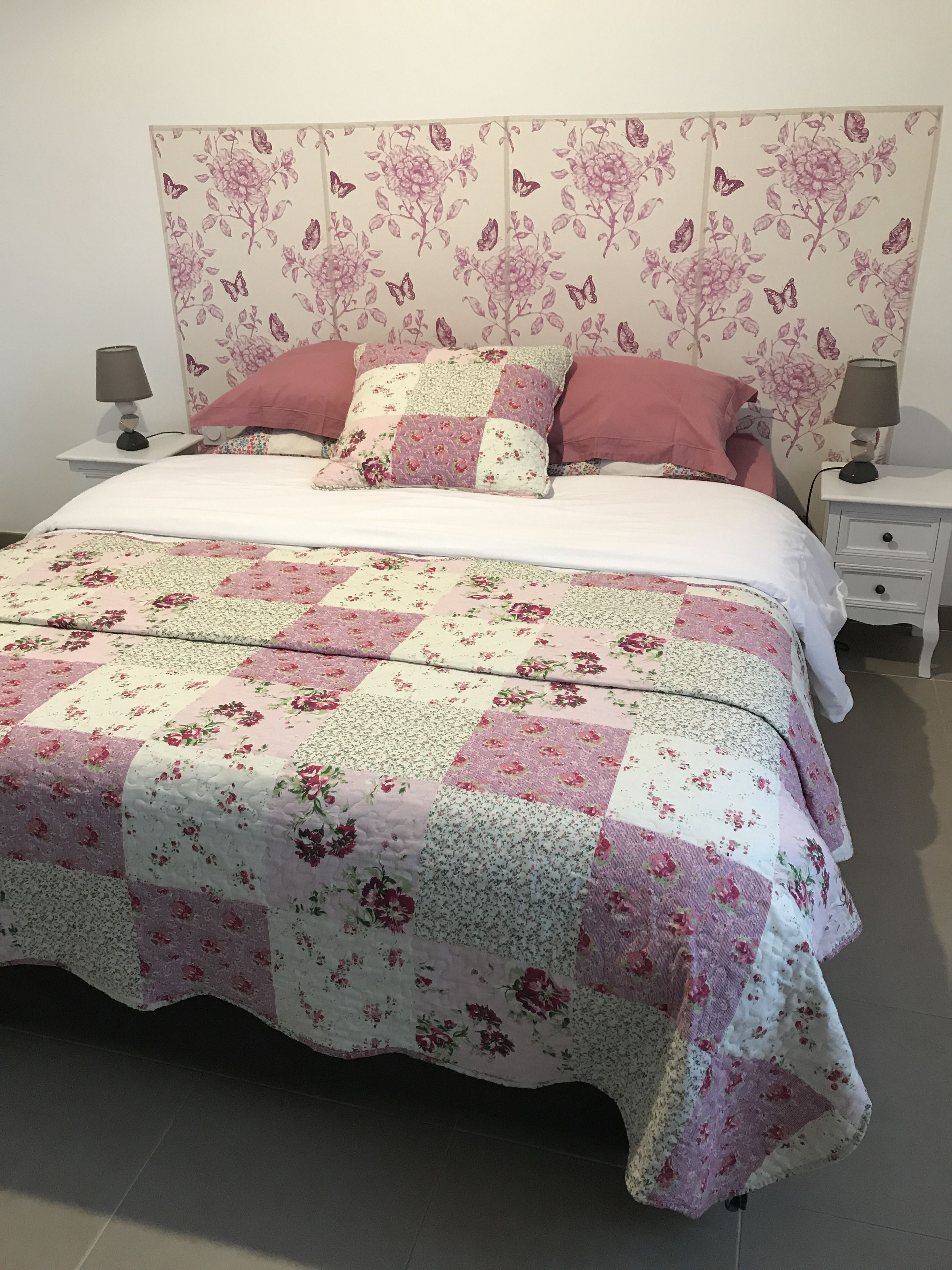 En pænt redt seng med et blomstret sengetæppe, to sengeborde med lamper, og en sengegavl med blomstermønster.
