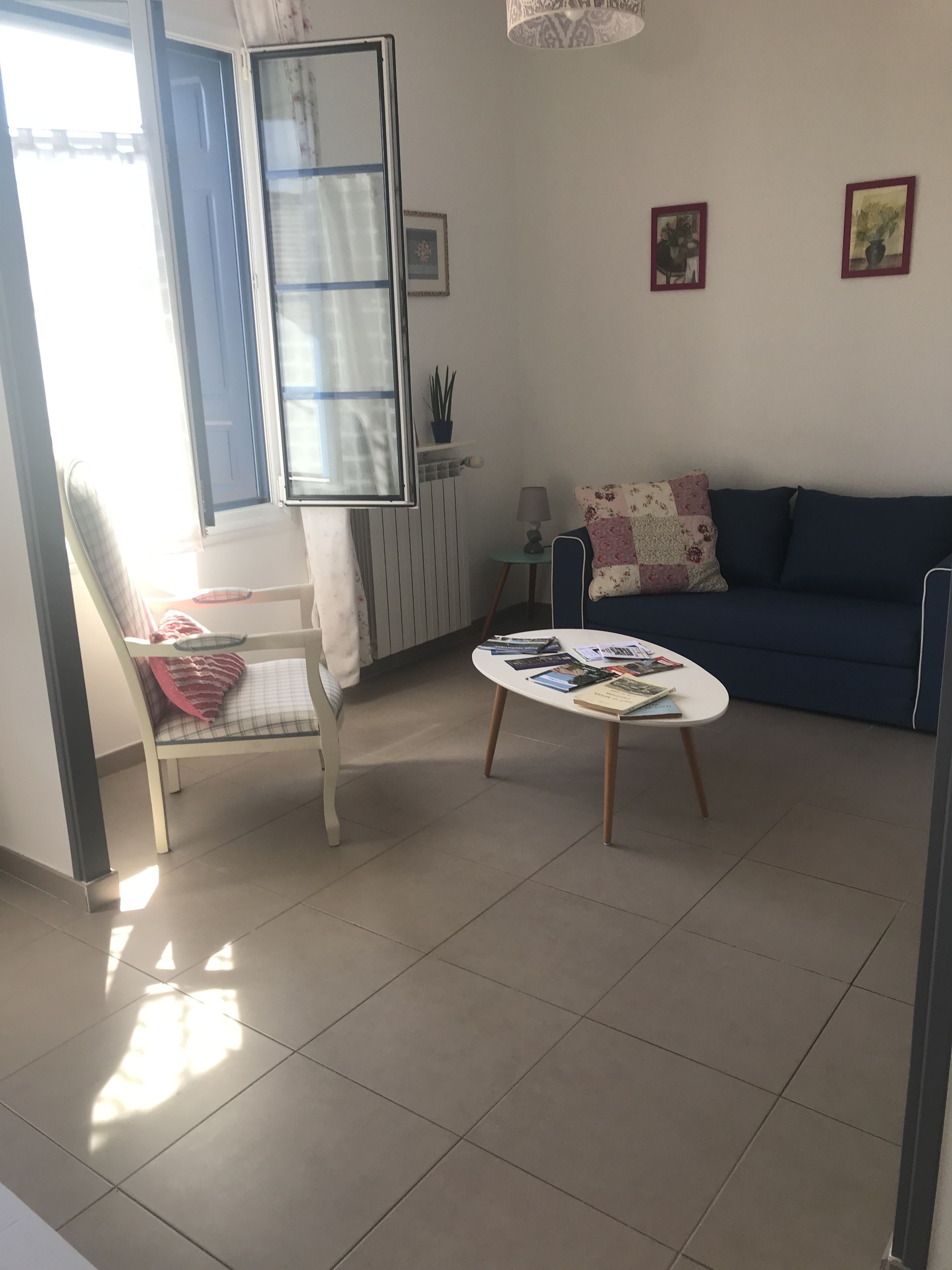 A living room with a sofa, a small table with books, a chair with a cushion, a lamp, and a window with blinds.