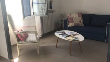 A living room with a sofa, a small table with books, a chair with a cushion, a lamp, and a window with blinds.