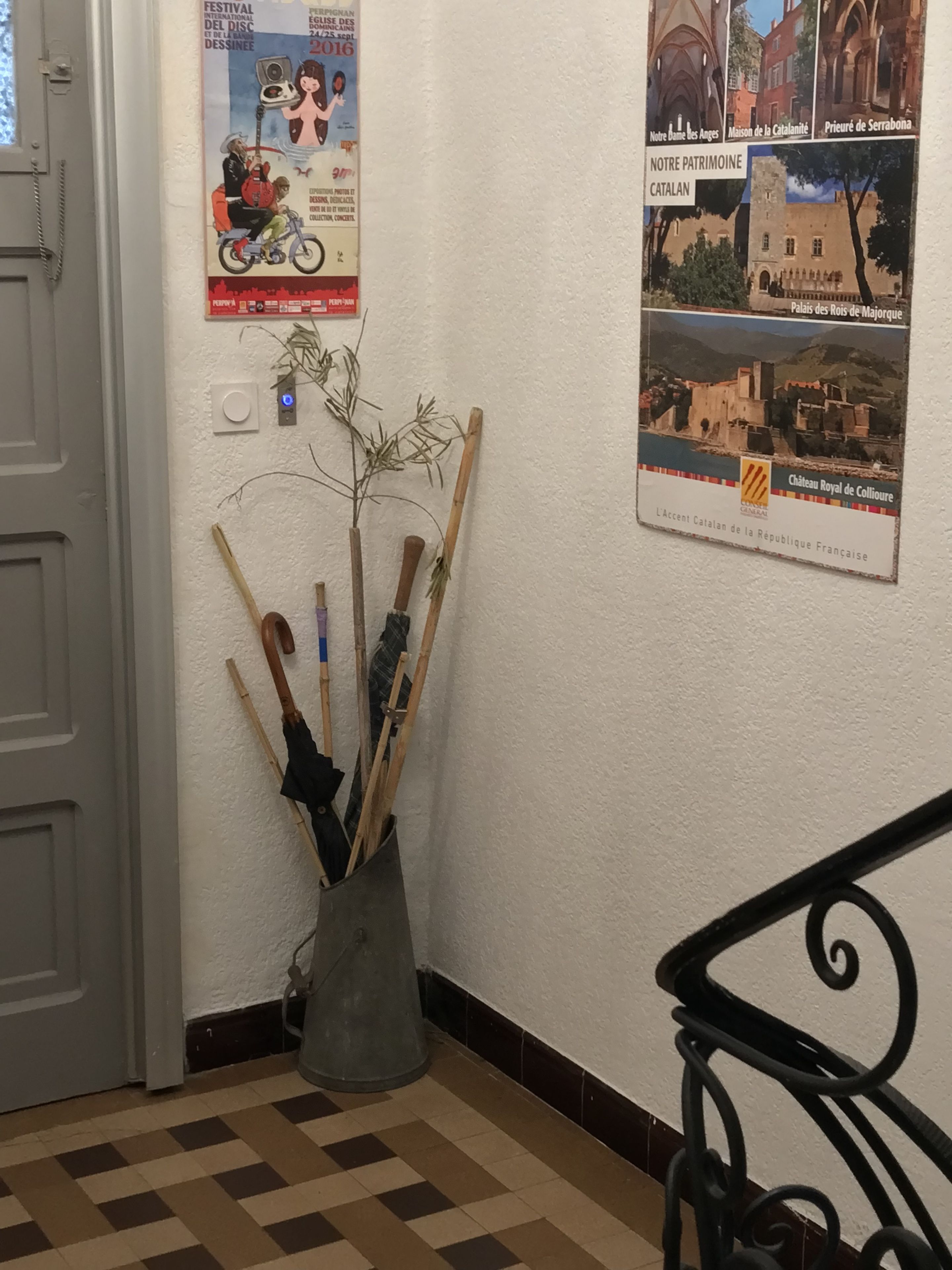 Ein Flur mit einer Tür, einem Festivalplakat, einem Metallbehälter mit Reinigungsausrüstung und einer Treppengeländer.