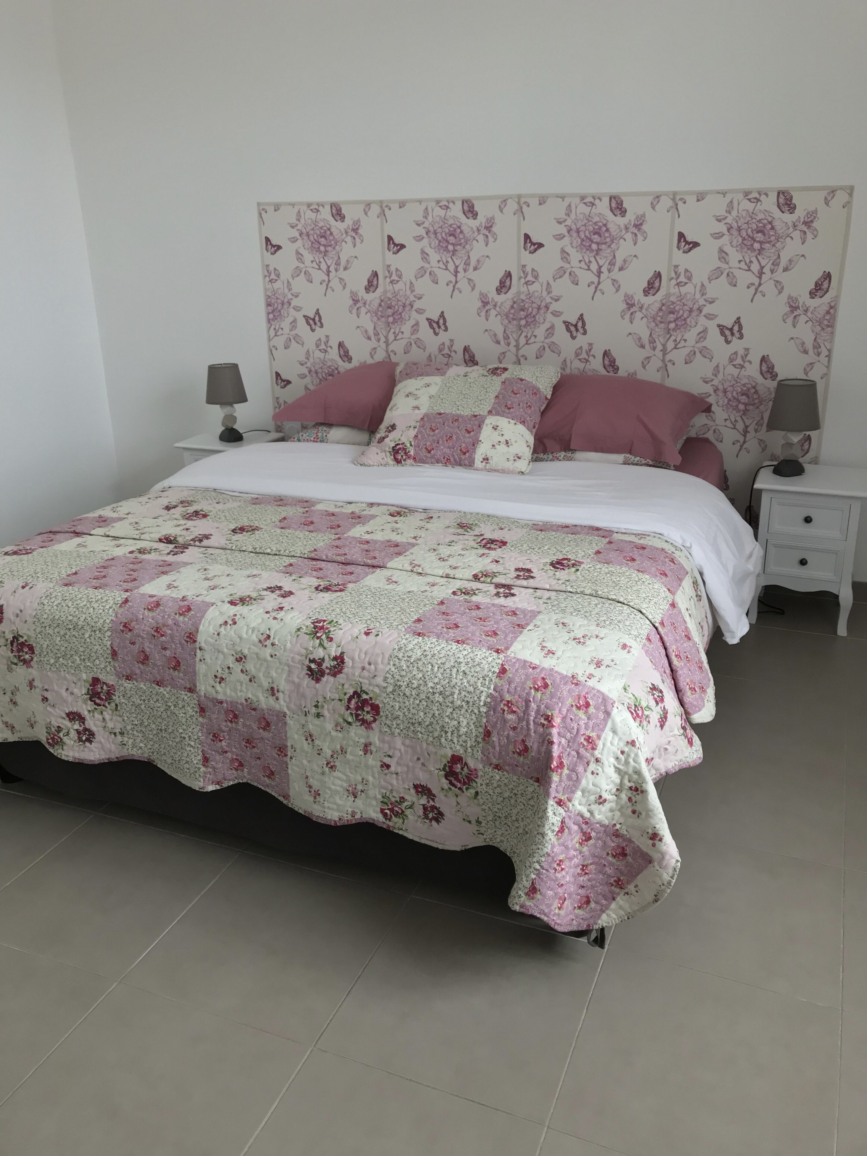 A neatly made bed with a floral quilt, a white headboard with a butterfly pattern, and a bedside table with a lamp.