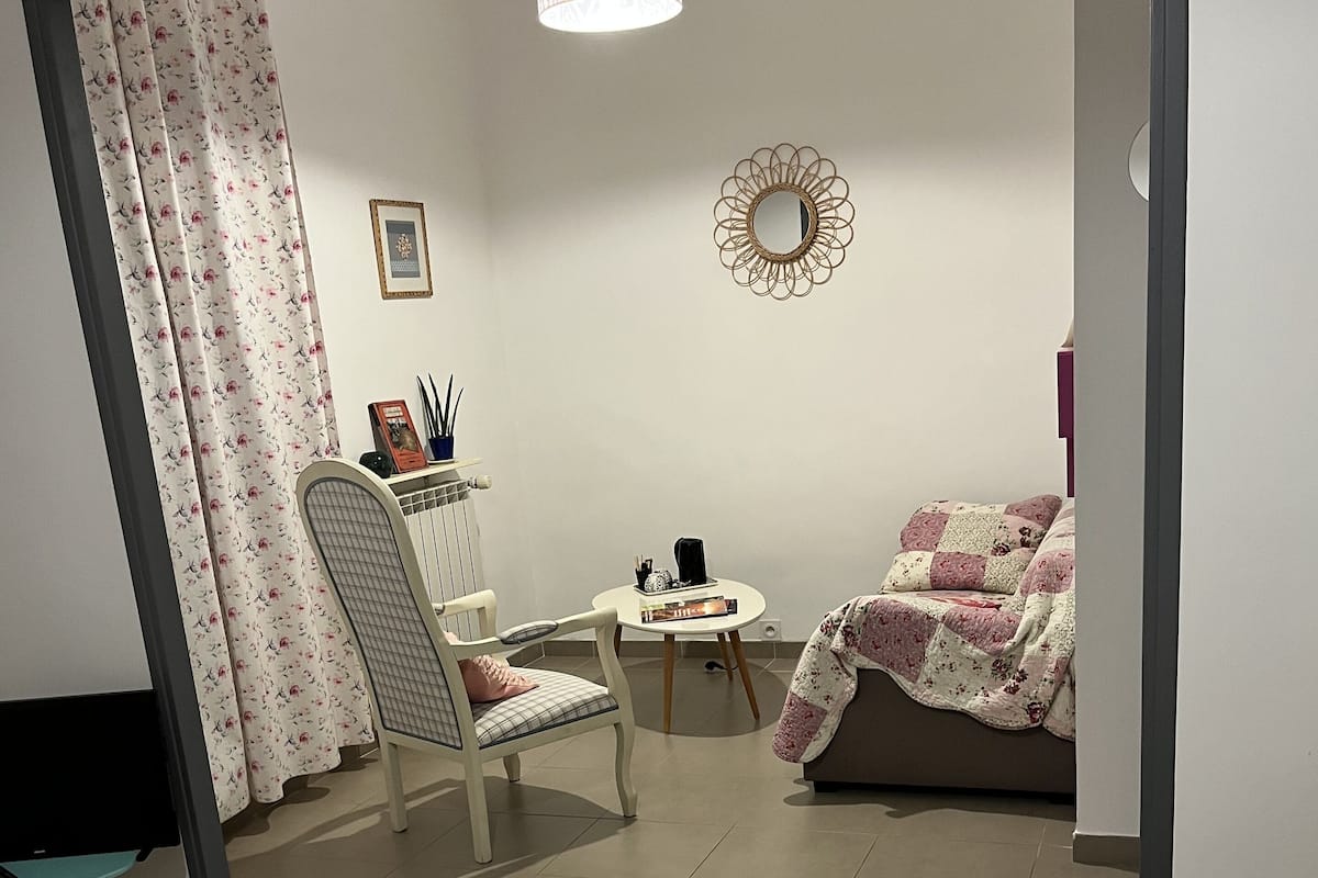 Un dormitorio con cama, una silla, una mesita, un sofá, un espejo y una cortina.