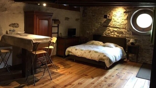A rustic bedroom with a wooden bed, a small desk with a chair, and a stone wall with a circular window. - Chambres d'Hôtes les Hirondelles (Folkling)