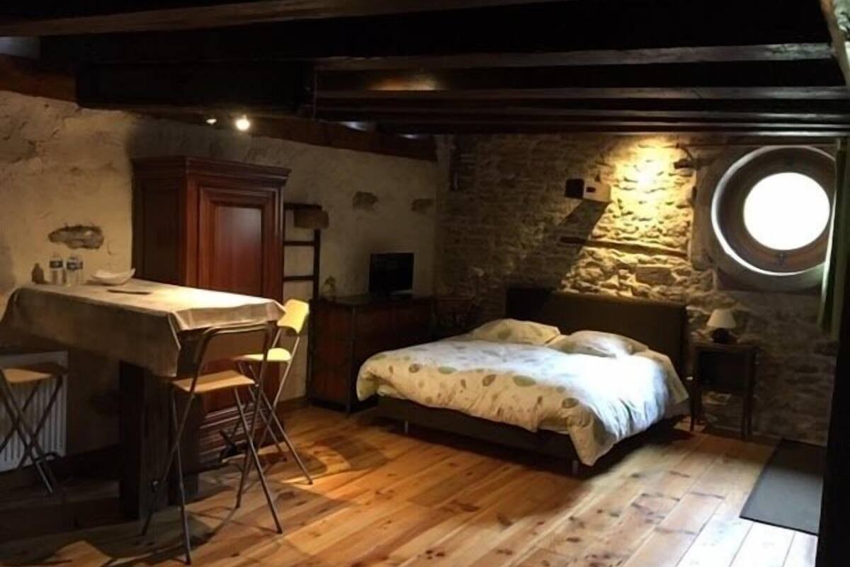 Une chambre à coucher rustique avec un lit en bois, un petit bureau avec une chaise et un mur en pierre agrémenté d’une fenêtre circulaire.