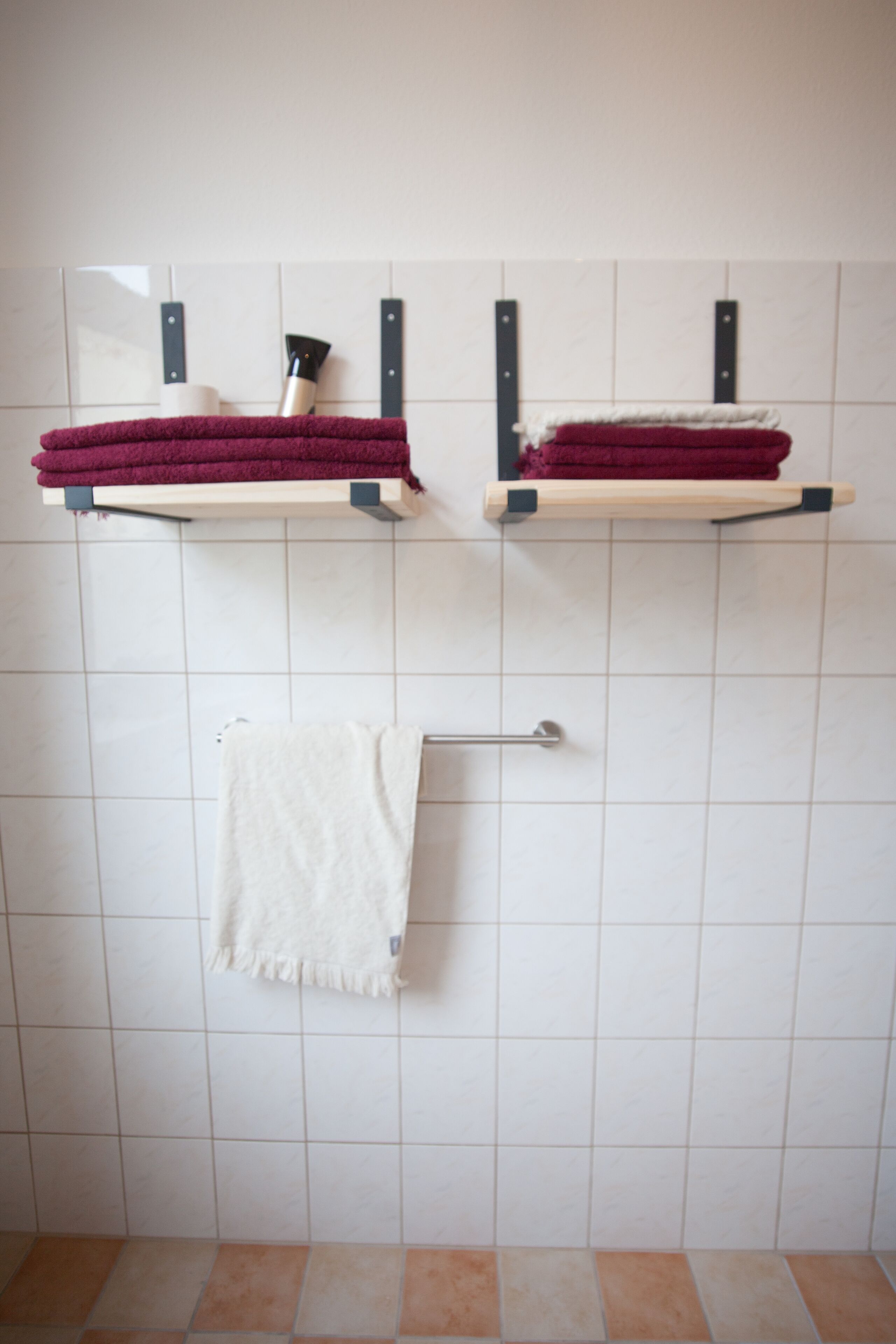A bathroom with white tiled walls, two towel racks with folded towels, and a metal towel rack with a single towel.