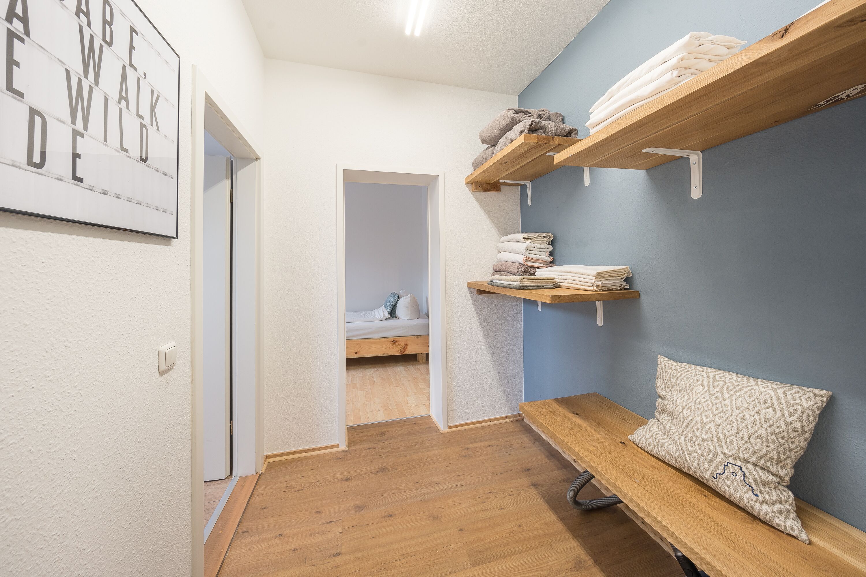 Un couloir avec un banc en bois, un lit à l’arrière-plan et une étagère fixée au mur où sont pliées des serviettes.