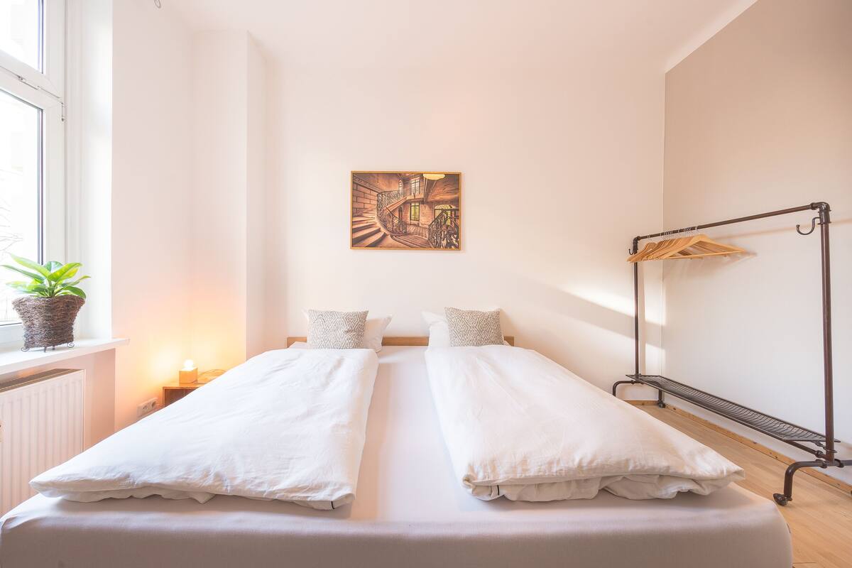 En dobbeltseng med hvide sengetøj, et natbord med lampe, et billedværk på væggen og en radiator.