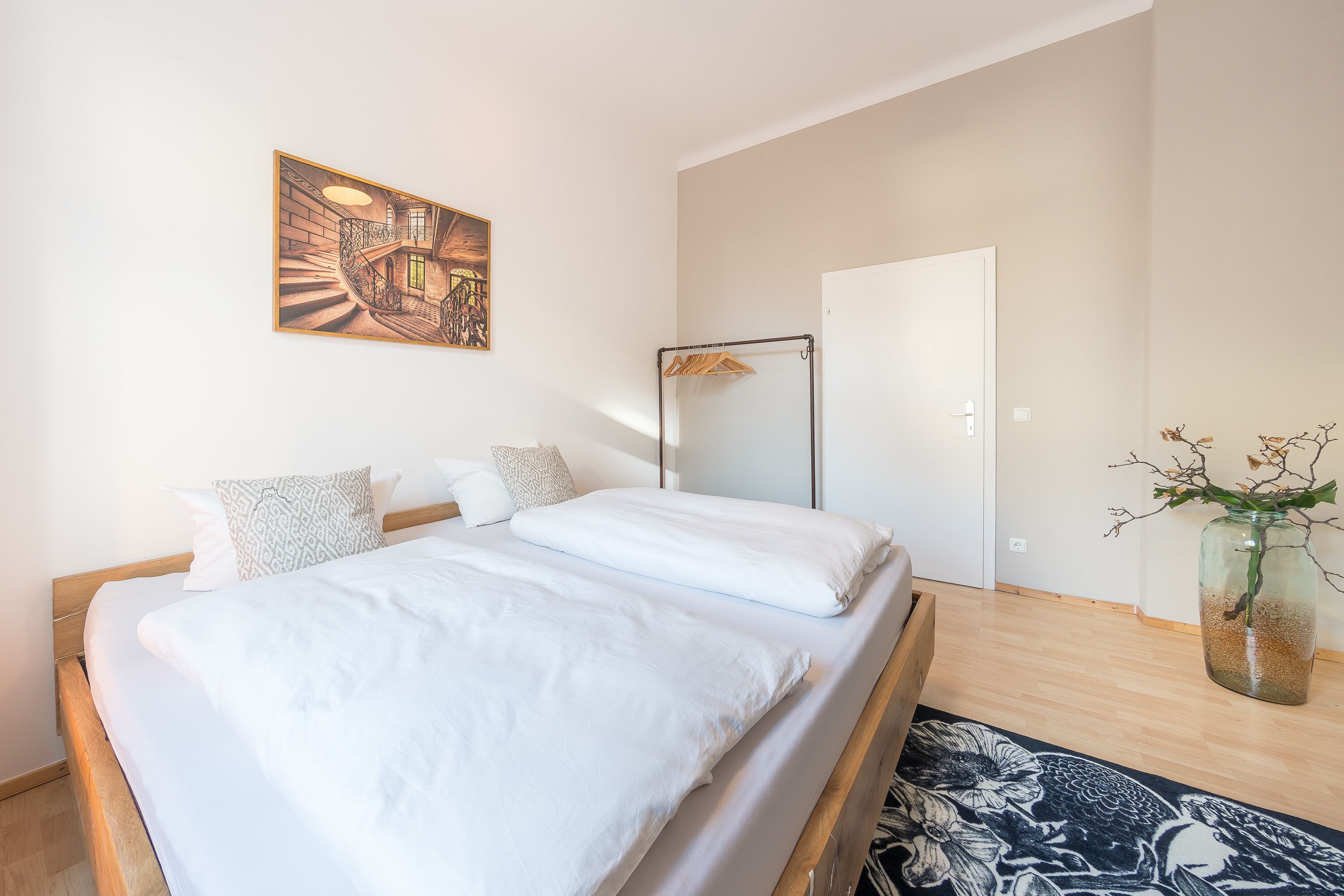 A bedroom with a bed, a wooden headboard, a painting, a floor lamp, a wardrobe, and a vase with branches.