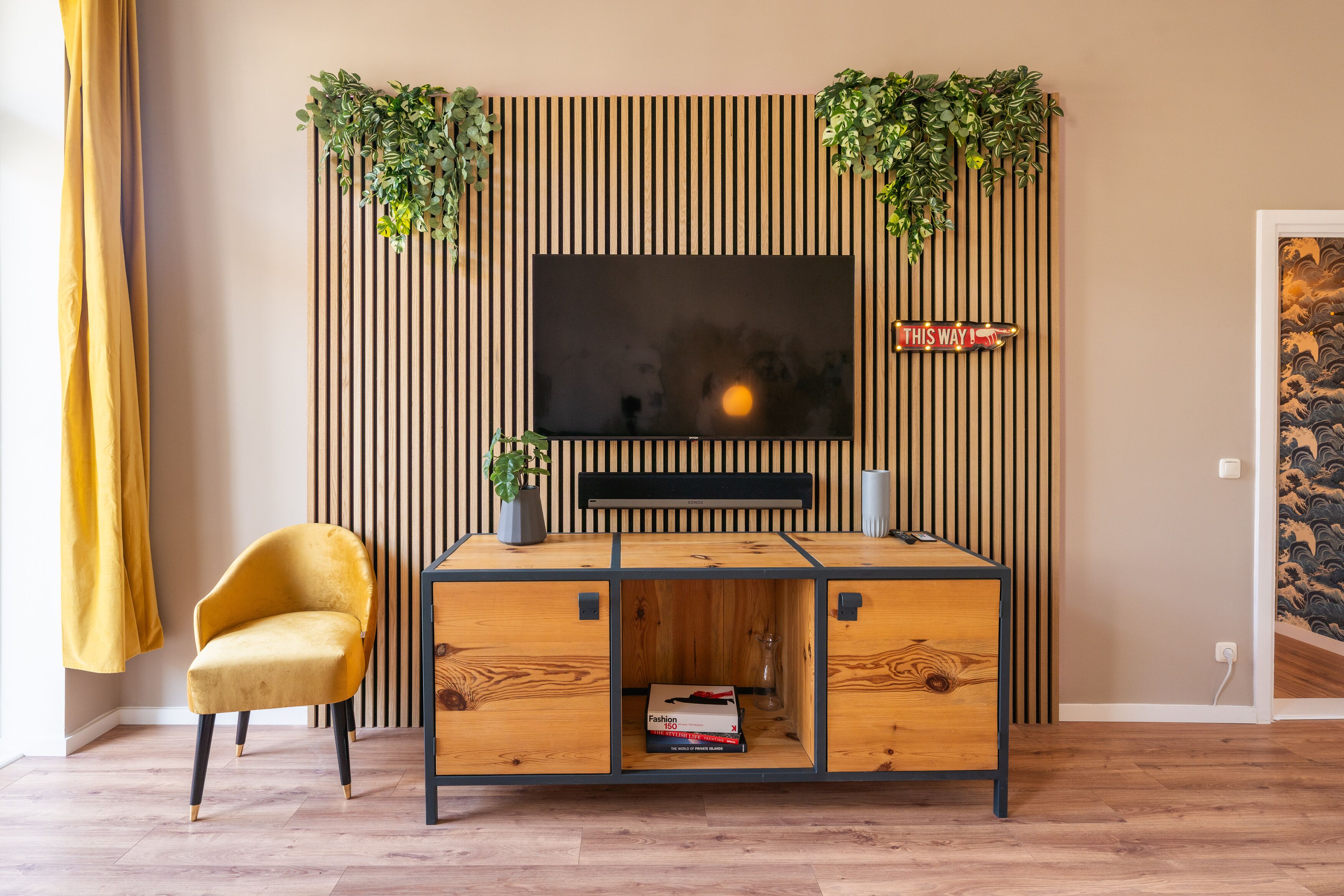 Un salon moderne avec un téléviseur à écran plat fixé à un mur en bois, un fauteuil jaune et un meuble de rangement en bois agrémenté d’un vase en verre et d’un livre.