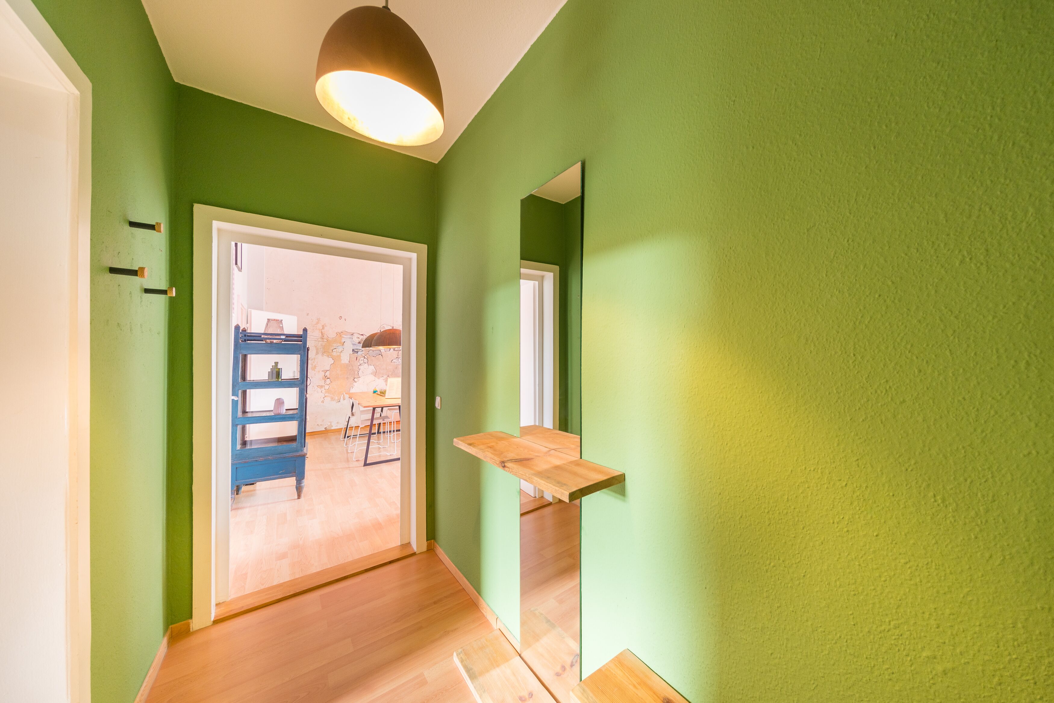 A hallway with green walls, wooden flooring, a blue shelf, and a hanging light fixture.