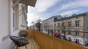 Ein Balkon mit zwei schwarzen Stühlen, Holzboden und Blick auf eine Stadtlandschaft mit Gebäuden und einer Straßenbahn.