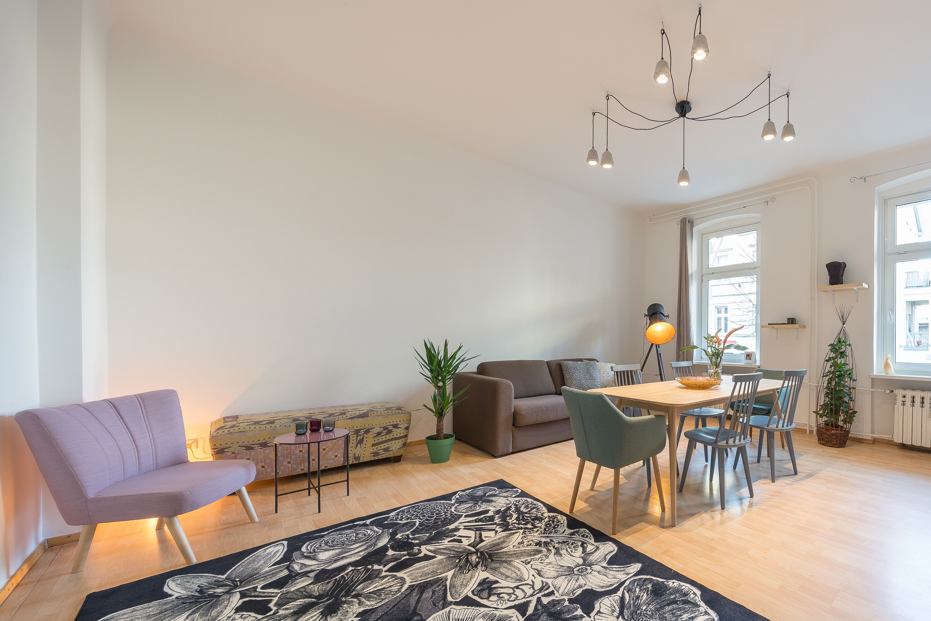 Una sala de estar moderna con un sofá, una mesa de comedor con sillas, una araña y ventanas amplias.