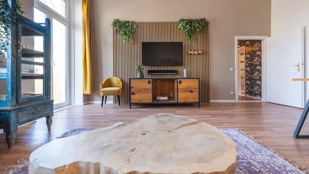 Ein Wohnzimmer mit einem großen Holztisch, einem Fernseher an einer Holzwandkonsole und einem gelben Sessel.