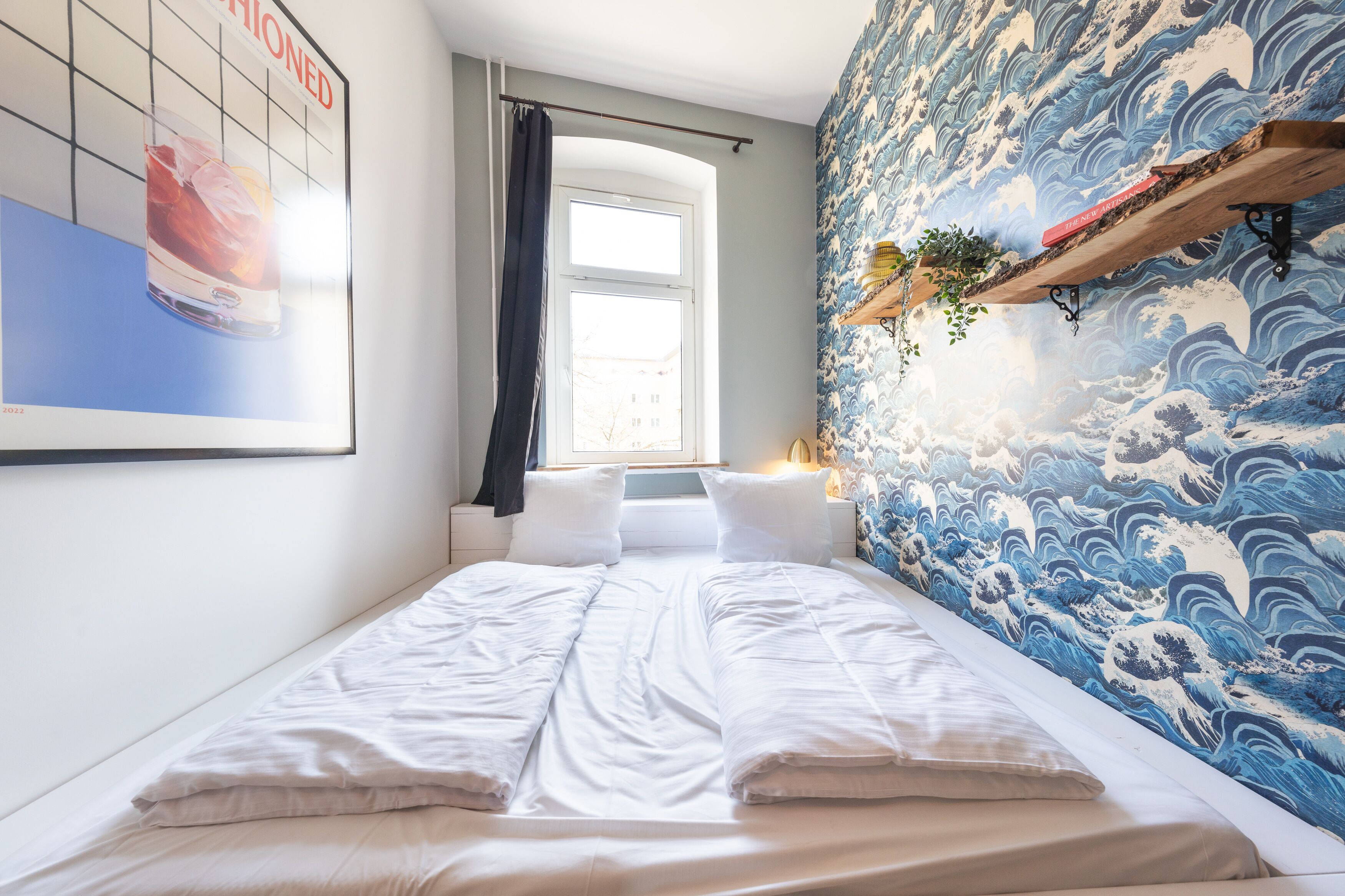 Un lit bien fait, avec du linge de lit blanc, une fenêtre avec un rideau et un mur orné d’un motif ondulé bleu.