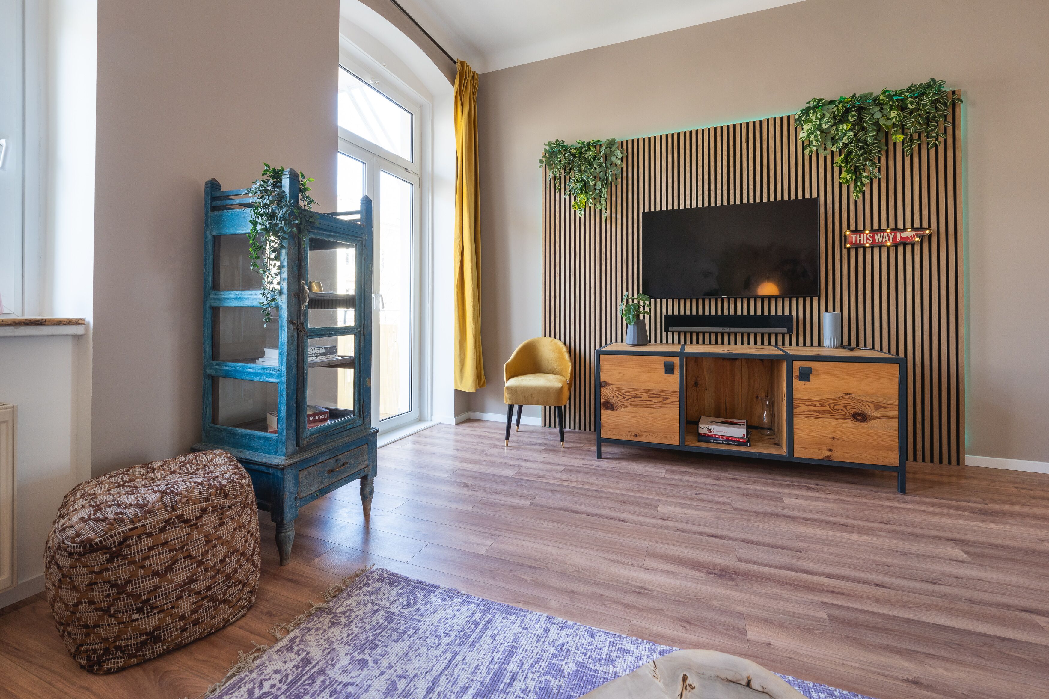 Un soggiorno con una TV fissata a una parete di legno, un mobiletto blu con ante in vetro, una poltrona gialla e un poggiapiedi decorato.