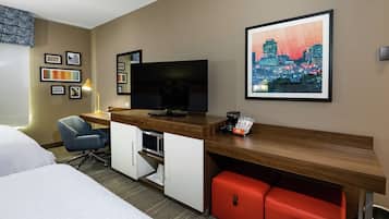 Hotellihuone, jossa on sänky, työpöytä tietokoneella, televisio ja punainen säilytysyksikkö.