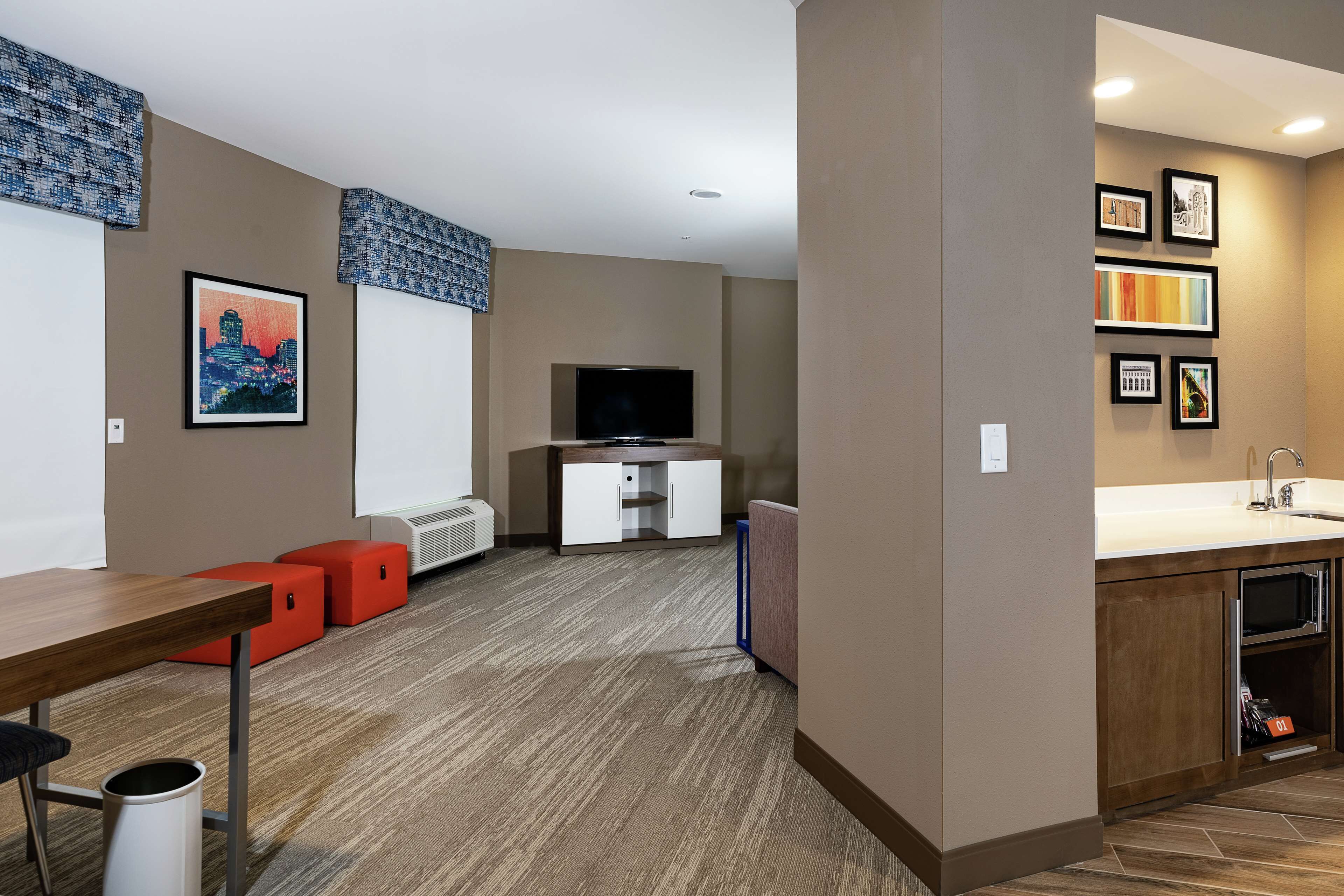 A modern hotel room with a flat-screen TV, a kitchenette, a dining area with a wooden table and red ottomans, and artwork on the walls.