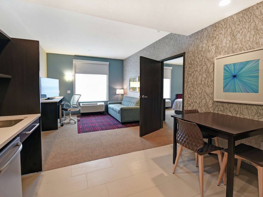 Una habitación de hotel moderna con kitchenette, un comedor con mesa y sillas, un sofá y un amplio ventanal.