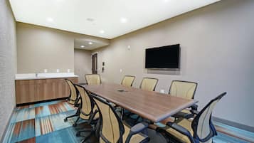 A conference room with a long table, chairs, a flat-screen TV, and a kitchenette area.
