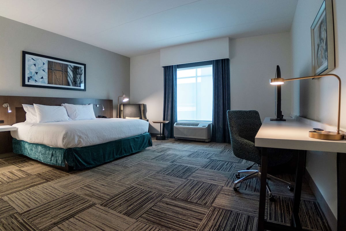 Een hotelkamer met een bed, bureau, stoel, airconditioning en een groot raam met gordijnen.