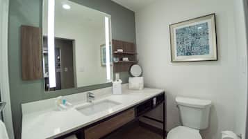 A modern bathroom with a large mirror, white sink, and toilet. There are wooden cabinets and a framed artwork on the wall.