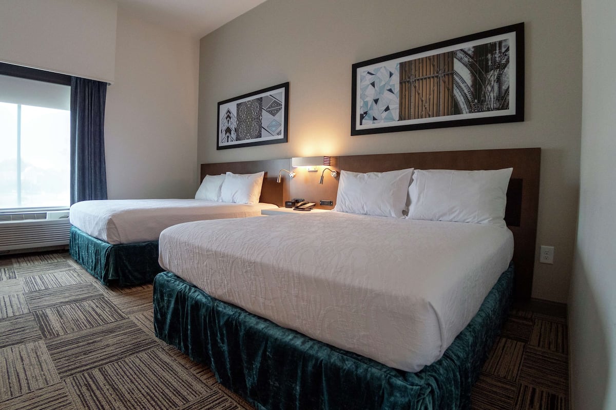 Een hotelkamer met twee bedden, een groot raam met gordijnen en twee ingelijste kunstwerken aan de muur.