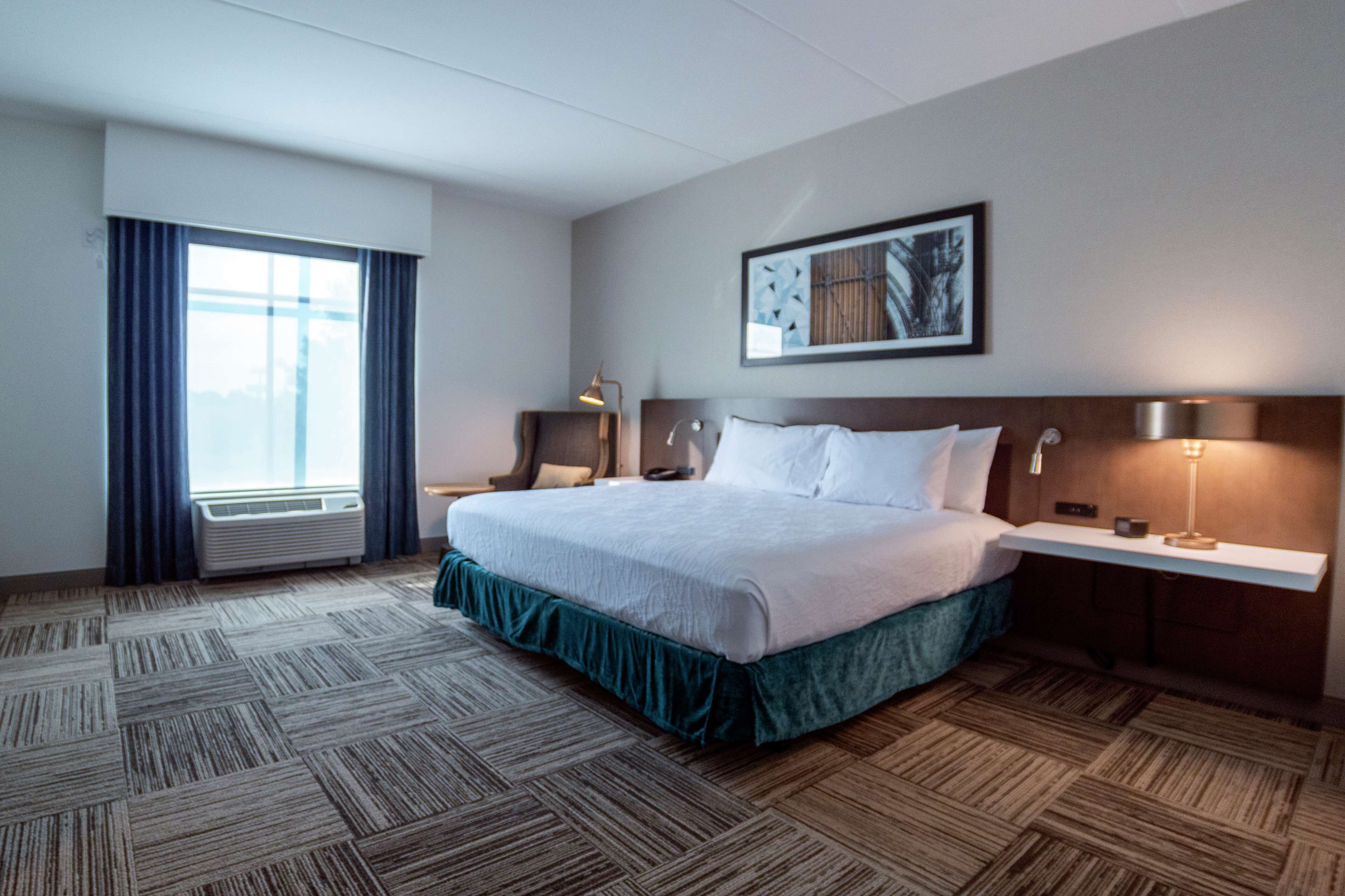 A hotel room with a large bed, a bedside table with a lamp, a window with curtains, and a wall-mounted picture.