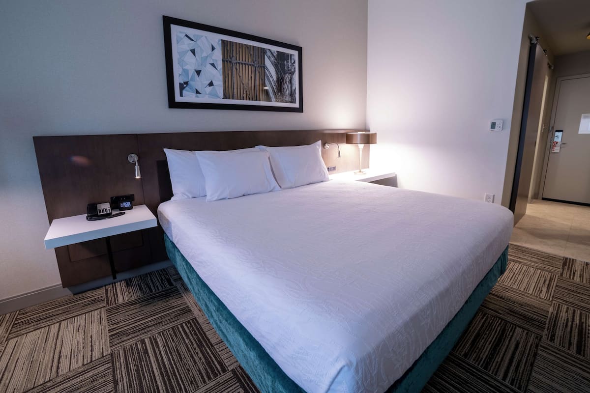 Een hotelkamer met een groot bed, een nachtkastje met een lamp, een lijst met een afbeelding aan de muur en een vloerbedekking.