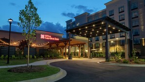 Ein Hotel mit Eingangsbereich, einem überdachten Gang, einer begrünten Anlage mit Bäumen und einer gut beleuchteten Fassade.