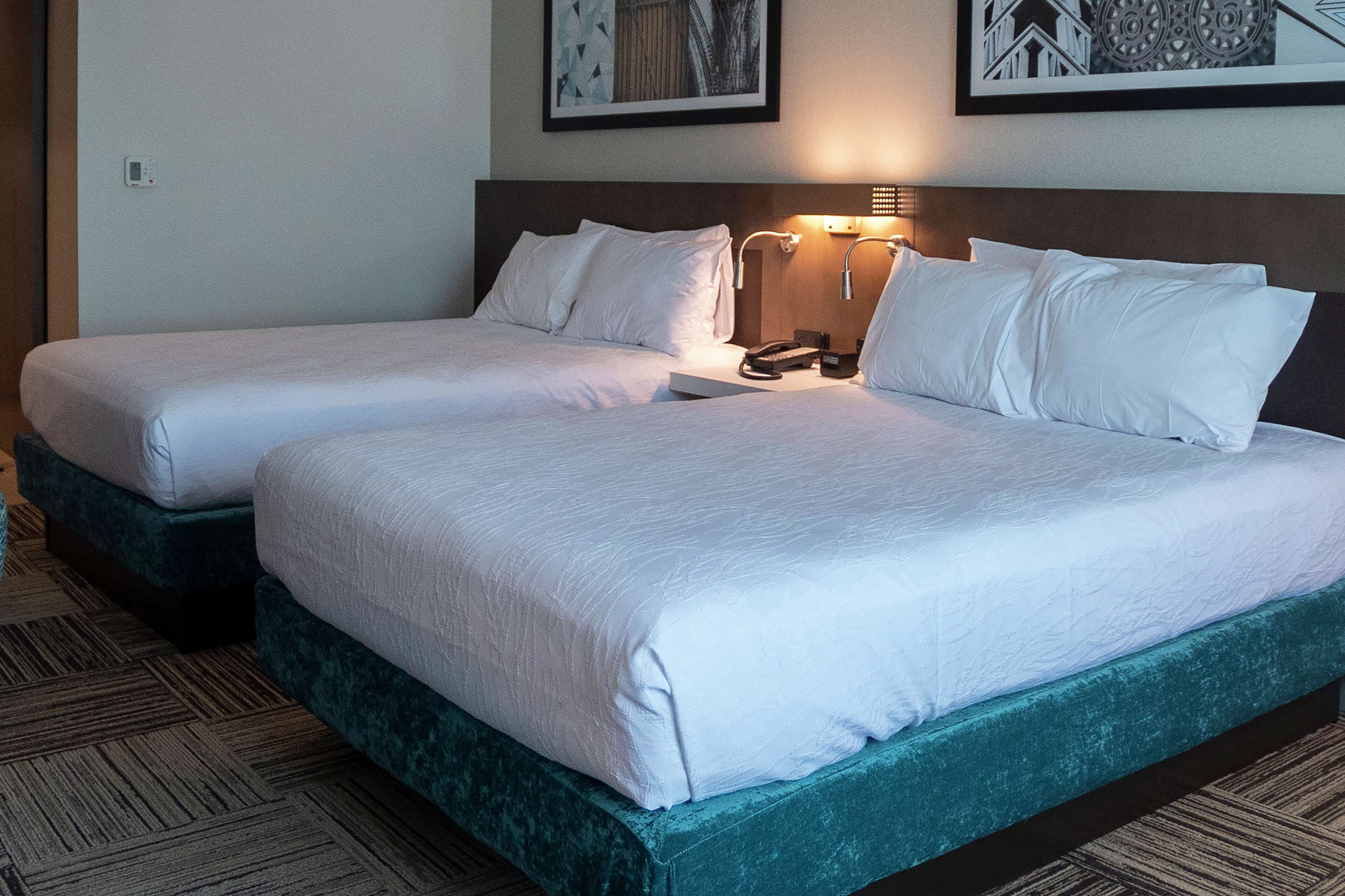 Habitación de hotel con dos camas, cada una con ropa blanca, cabeceros, lámparas de noche y piso alfombrado.