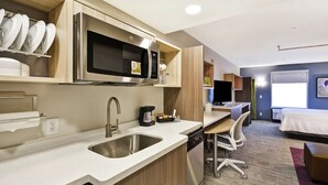 A modern hotel room with a kitchenette, including a microwave, sink, and coffee maker. There is a desk with a chair, a bed, and a window with a view. - Home2 Suites by Hilton Warner Robins (Warner Robins)