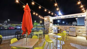 Un patio al aire libre con sillas y mesas amarillas, luces colgantes y una pared de piedra con una parrilla.