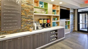 A hotel reception area with a stone wall, a sign stating 'HOME GUARANTEE', a coffee station, and a TV mounted on the wall. - Home2 Suites by Hilton Warner Robins (Warner Robins)
