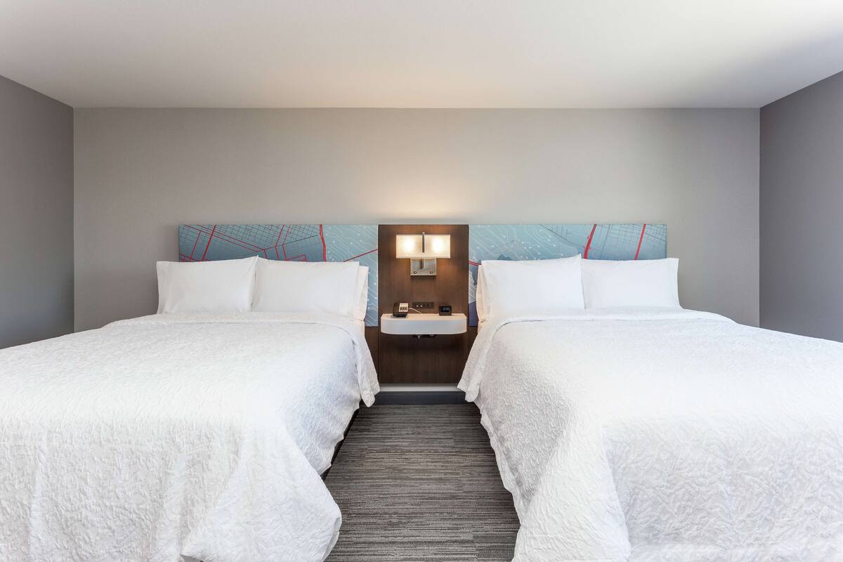 Habitación de hotel con dos camas, una mesita de noche, una lámpara de pared y un lavamanos pequeño.