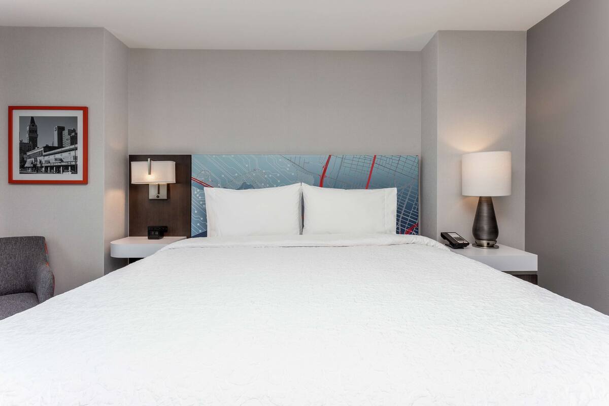 Habitación de hotel con una cama grande, dos mesitas de noche, una lámpara, una silla y un cuadro en la pared.