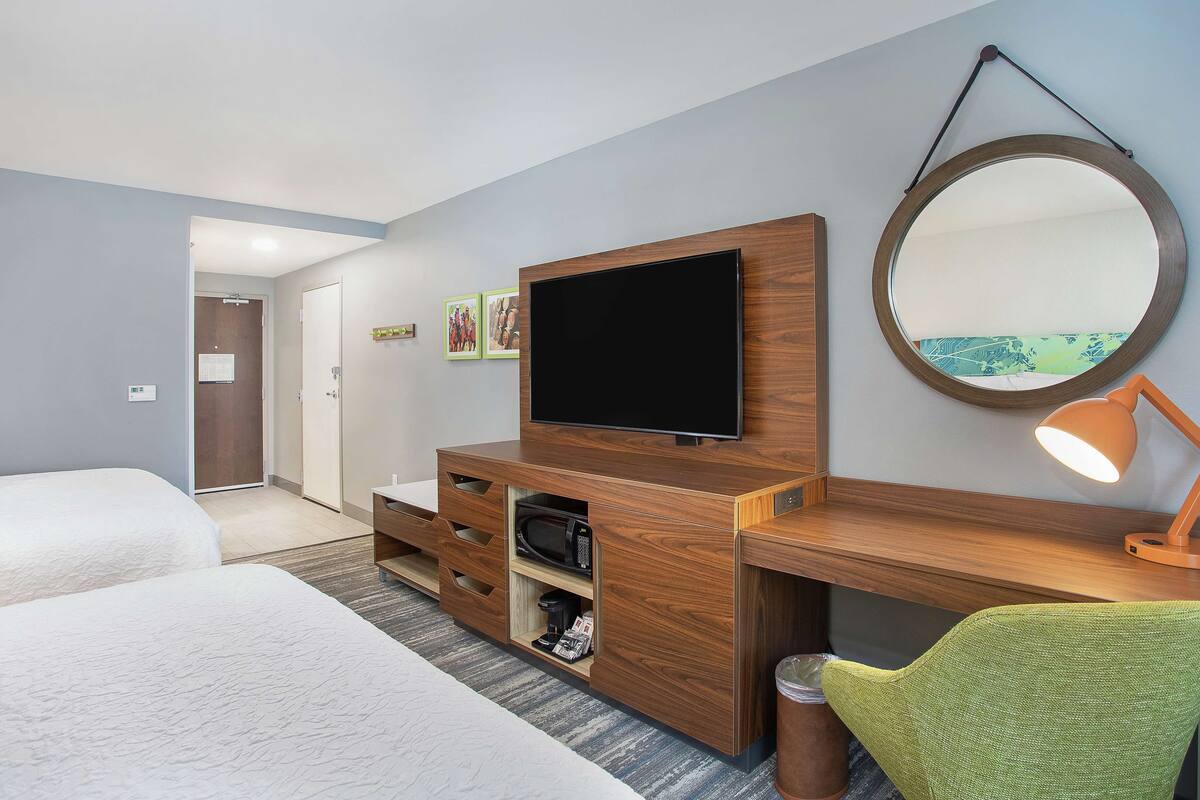 A hotel room with a wooden desk, a flat-screen TV, a green chair, and a large mirror.