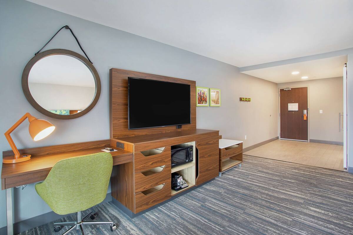 A hotel room with a wooden desk, a green chair, a flat-screen TV, a round mirror, and a small shelf.