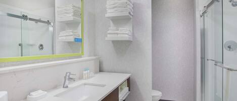 Un baño moderno con cabina de ducha de vidrio, un lavabo blanco con espejo y toallas ordenadas en estantes.