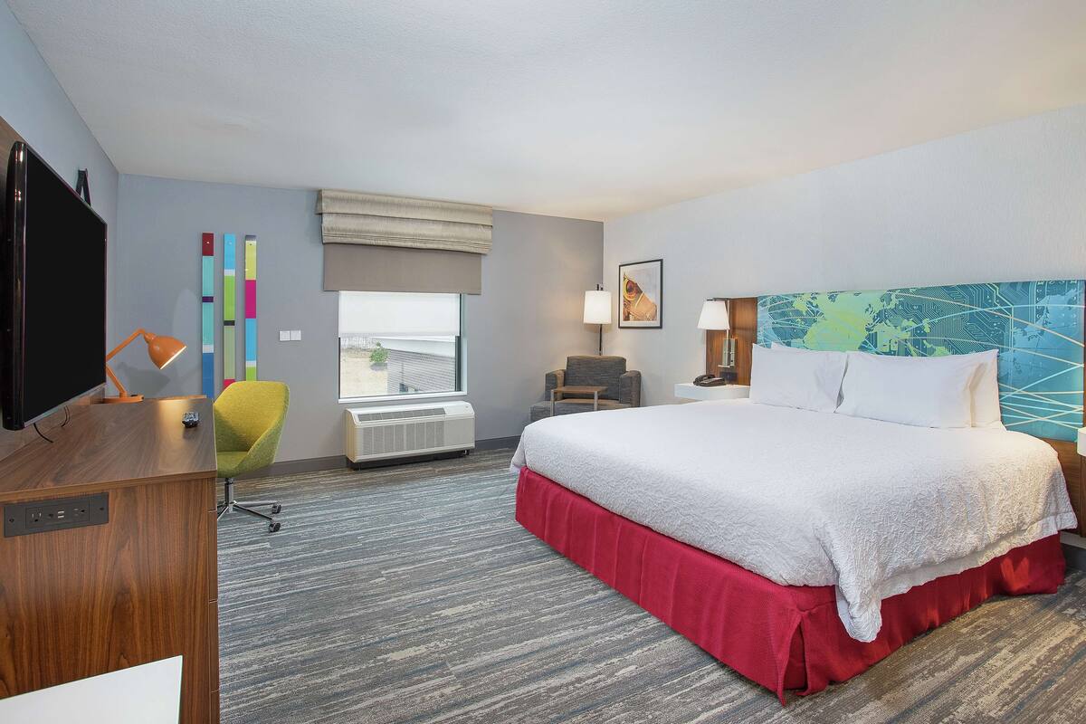 A hotel room with a large bed, a desk with a TV, a chair, a window, and a wall with colorful vertical elements.