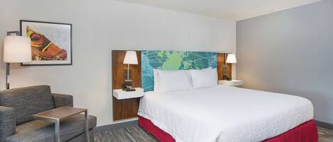 Quarto de hotel com cama, sofá, mesa pequena, mesa de cabeceira e abajur.
