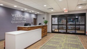 A reception area with a "Welcome to the Blue Grass State" sign, a reception desk, a potted plant, and a view through the glass doors. - Hampton Inn Simpsonville (Simpsonville)