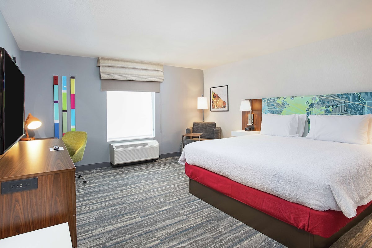 A hotel room with a bed, a desk with a lamp, a chair, a TV, and a window with a view.