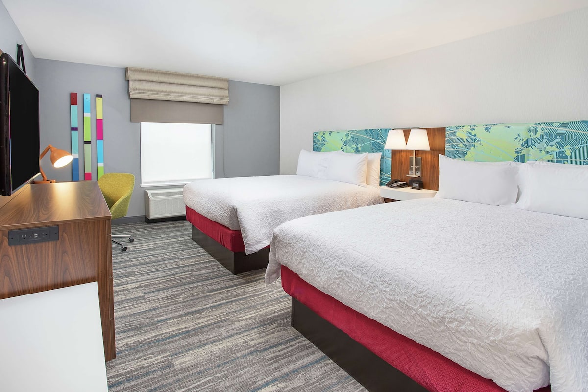 A hotel room with two beds, a desk, a chair, a TV, and a window with a view.