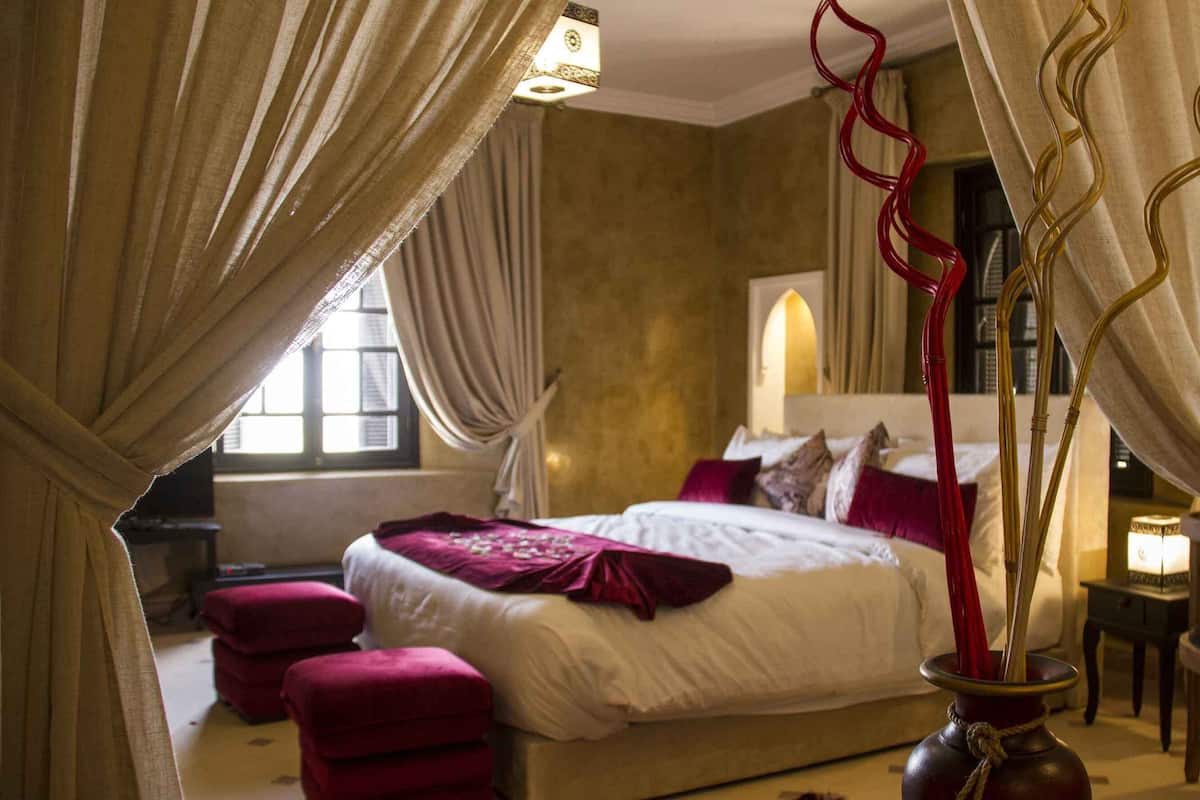 A bedroom with a bed, red ottomans, a vase with red branches, and curtains.