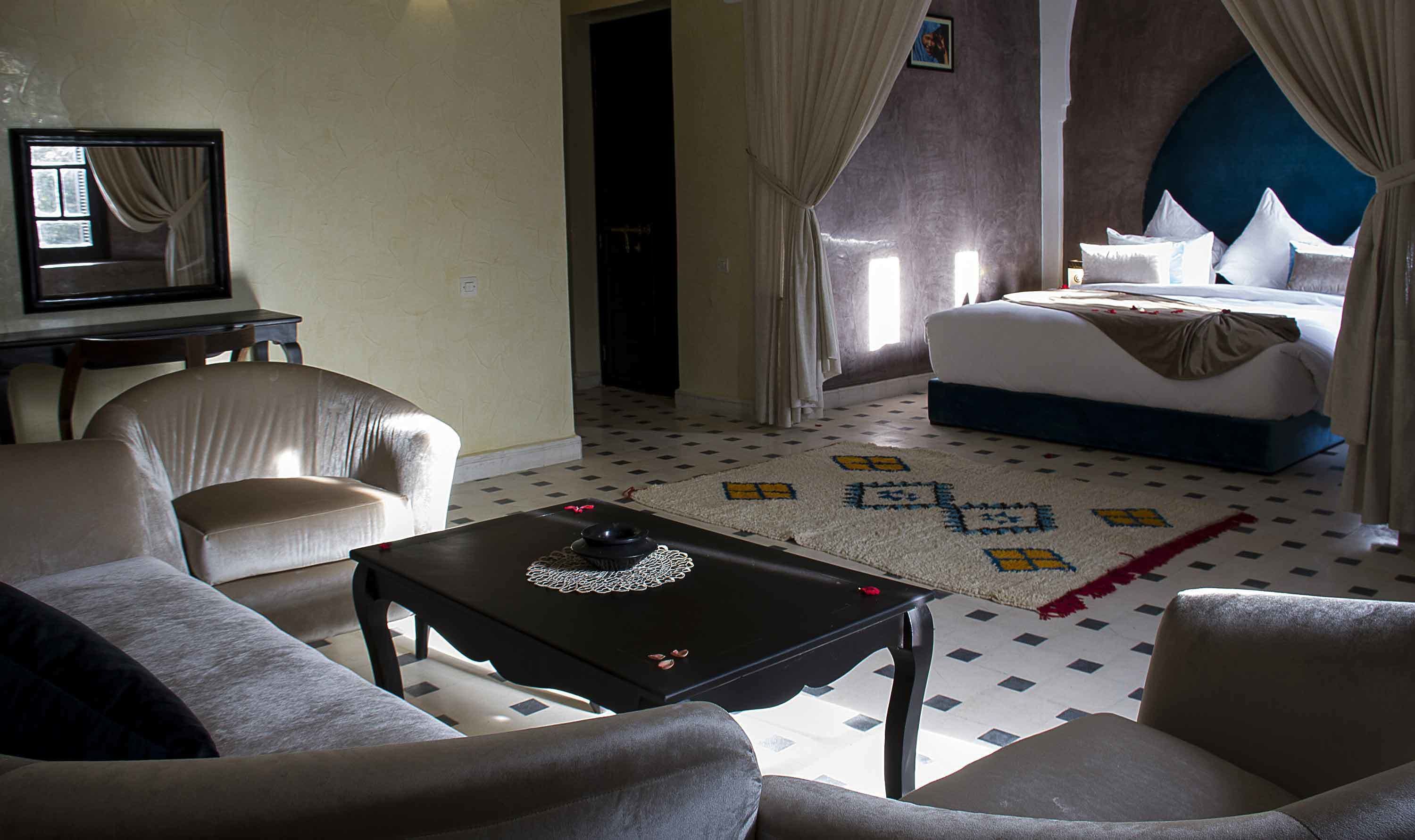 Habitación de hotel con cama, dos sillones, una mesita con un objeto decorativo, un tapete estampado y un espejo en la pared.