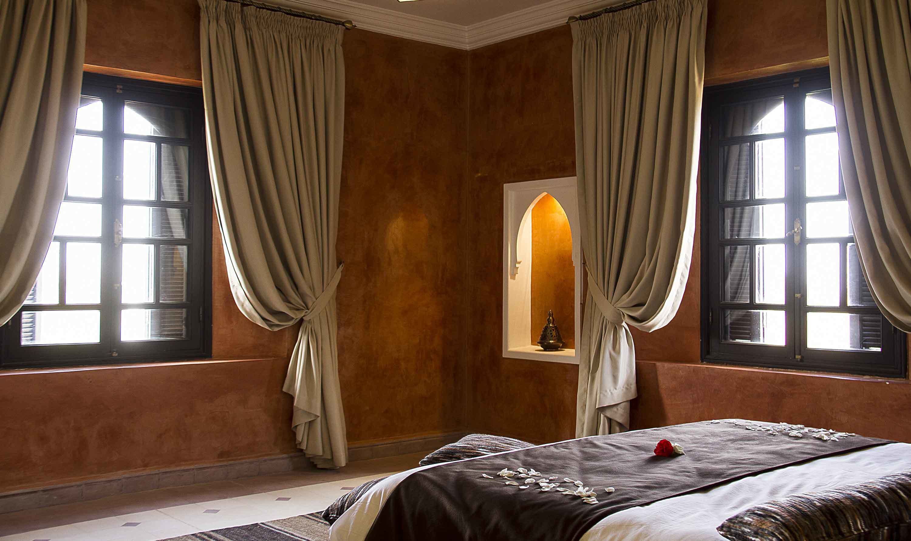 Ein Schlafzimmer mit zwei Fenstern, beigen Vorhängen, einem Bett mit dunkler Bettdecke und einem kleinen Spiegel über dem Bett.
