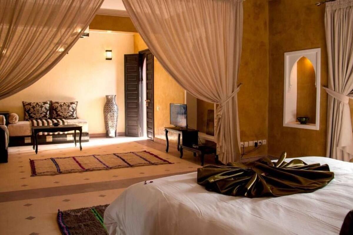 A hotel room with a bed, a TV, a desk, and a chair. There is a window with curtains, a vase on the desk, and a rug on the floor.
