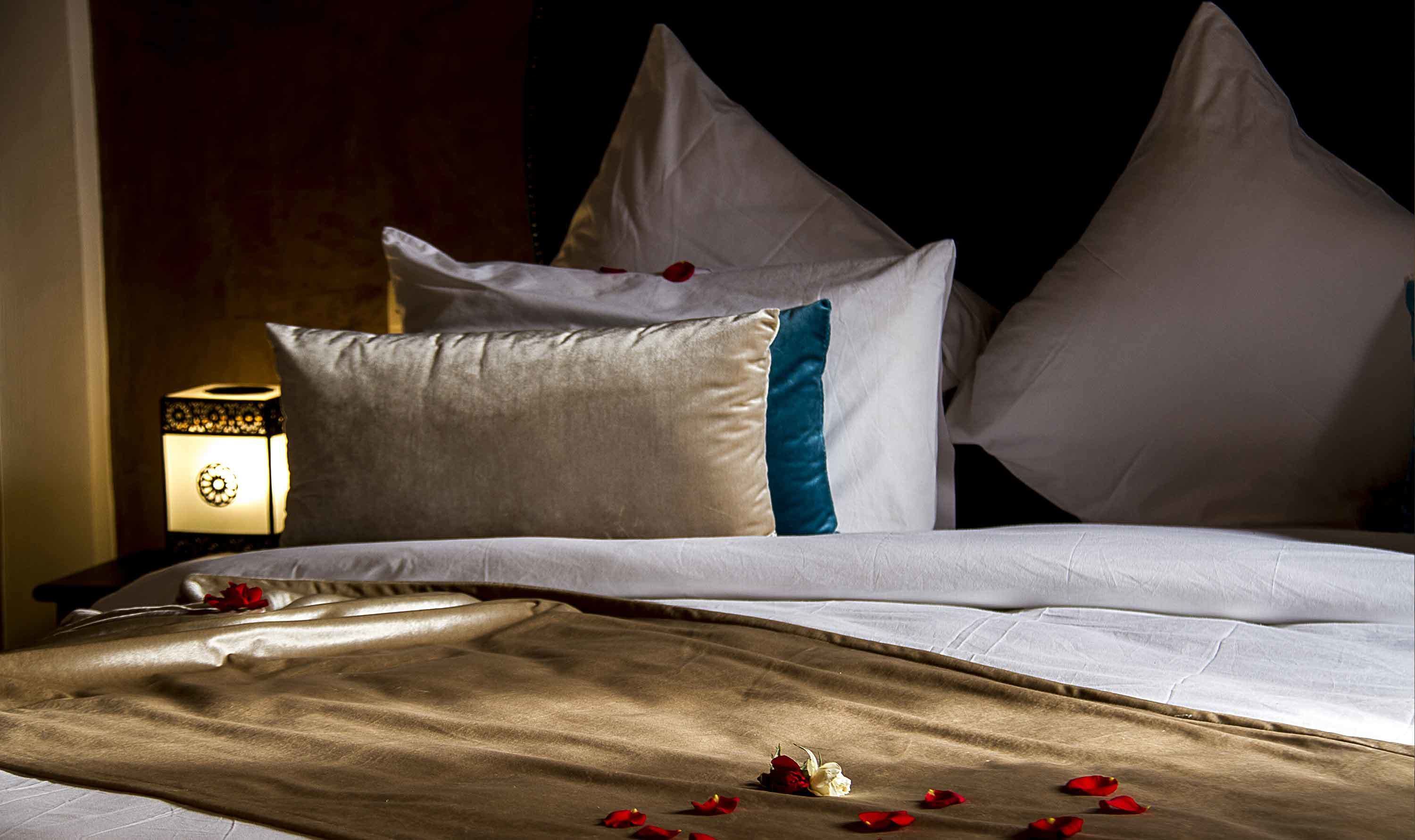 A neatly made bed with white and blue pillows, a beige bedspread, and scattered rose petals.