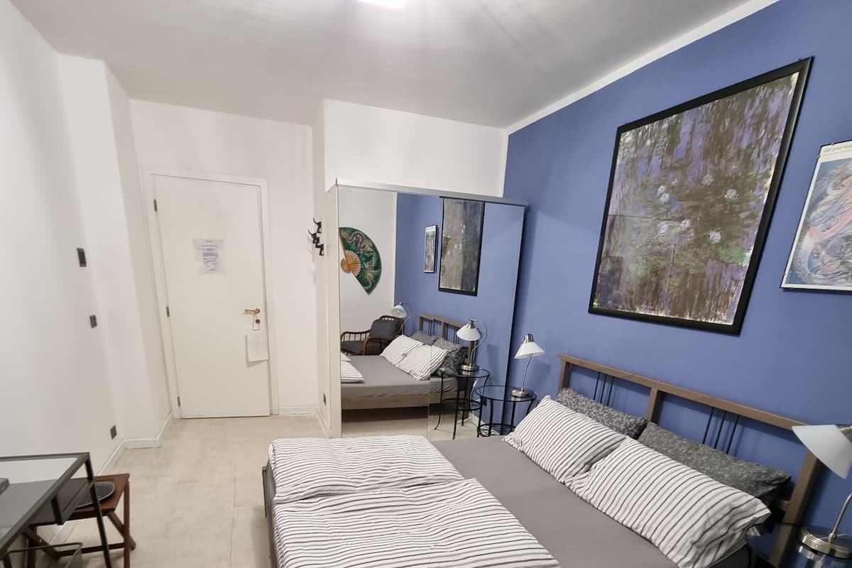 Un dormitorio con cama, escritorio y silla. Se ve una puerta y una pared con un cuadro.