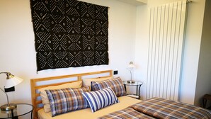 Un lit double avec une couette à carreaux, une table de chevet avec une lampe, une tapisserie à motifs et une fenêtre avec des stores.